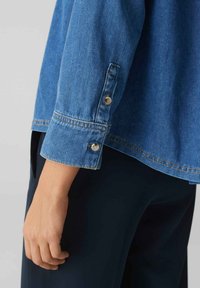 Camicia in denim blu con vestibilità rilassata, caratterizzata da polsini con bottoni, cuciture a contrasto e una texture liscia. Posizionata accanto a pantaloni scuri.