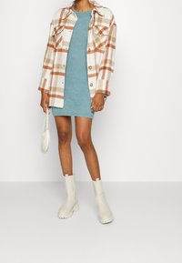 Beige och brun rutig shacket över en turkos stickad klänning med en fluffig textur. Ett par vita ankelskor och en liten vit handväska.