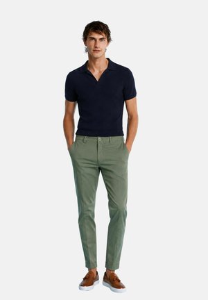 Uomo in piedi con le mani in tasca, indossa una polo blu navy a maniche corte, pantaloni verdi e mocassini marroni con suola bianca.