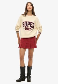 Cremefarvet sweater med "SUPER DRY" tekst, parret med en rød fløjlskørt med folder; fuldendt med sorte knæhøje støvler.