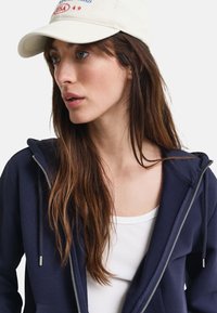 Marine blouson met rits en trekkoorden, gedragen over een witte tanktop. Beige pet met rode en blauwe tekst. Lange, kastanjebruine haren gedeeltelijk zichtbaar.