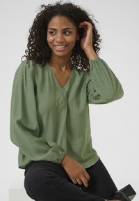 Lange mouwen, v-hals blouse in lichtgroen, gemaakt van textuurstof met gathering aan de manchetten. Gepaard met zwarte broeken en hakken.