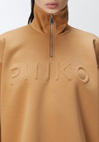 Felpa beige con colletto alto, chiusura a mezza zip e lettering "PINKO" in rilievo. Tessuto morbido e design semplice si concentrano sul comfort.