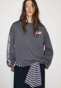 Szürke hosszú ujjú ing, bal ujján rózsaszín felirattal: "Merj kitűnni". Fekete nadrággal és derékon megkötött csíkos anyaggal párosítva.