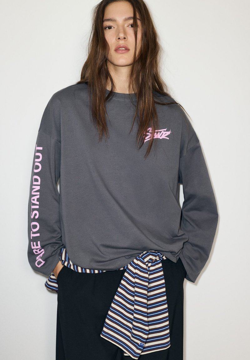 Szürke hosszú ujjú ing, bal ujján rózsaszín felirattal: "Merj kitűnni". Fekete nadrággal és derékon megkötött csíkos anyaggal párosítva.