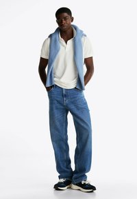 Camisa polo blanca, suéter de punto azul claro colocado sobre los hombros, jeans de mezclilla azules y zapatillas negras con cordones blancos.