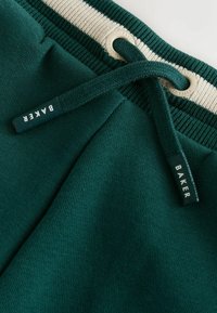 Pantalones deportivos de un verde oscuro con una cinturilla elástica gruesa que presenta una franaja crema. Incluyen un cordón y una etiqueta que dice "BAKER." Textura suave.