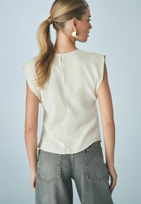 Blusa sin mangas color crema con costura trasera, combinada con jeans grises de tiro alto. Tejido suave, corte relajado y sutiles detalles en el dobladillo curvado.