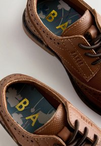 Hnedé kožené topánky s textúrovaným zvrškom s perforáciami. Vnútorná stielka má kvetinový vzor so zlatým nápisom "BAKER." Detail tmavej podrážky.