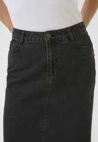 Jupe en denim noir à taille haute, avec une fermeture à boutons sur le devant, des poches latérales et une coupe slim. Texture lisse et forme structurée.