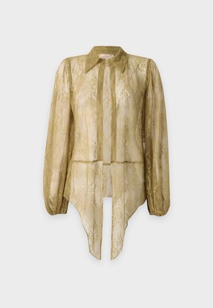 Femme portant un pantalon en dentelle olive transparente et un haut à manches longues assorti noué à la taille sur un haut de bikini avec des sandales blanches à talons.
