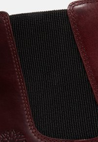 Gros plan d'un panneau en tissu élastique noir encadré de cuir cousu rouge foncé avec motif décoratif embossé sur l'angle en cuir.