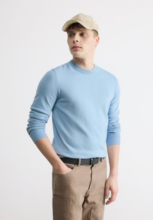 Giovane uomo con maglione azzurro chiaro, pantaloni beige con tasche e cappellino beige, in piedi con una mano in tasca, guarda verso sinistra.