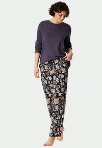 Kvinne iført en langermet lilla topp og svarte bukser med blomsterdesign, stående barbent mot en ensfarget bakgrunn.