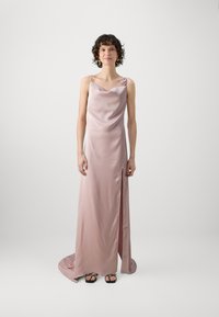 Lång, flödig klänning i mjuk pastellrosa satin med cowl-urringning, slits på sidan och en blommig detalj på ena axeln. Platta sandaler bärs nedanför.