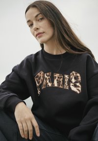 Melna kapuce ar vārdu "PARIJS" tekstūrētā brūn ā rakstā. Modelis sēž, demonstrējot neformālu, relaksētu piemērošanu.