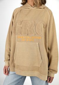 Beige Hoodie mit strukturiertem "GRC" Schriftzug, orangefarbenem "STUDIO COLLECTION" Text und einer Fronttasche. Besitzt einen lässigen Schnitt und gerippte Bündchen.