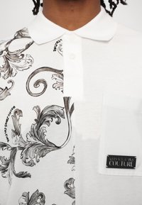 Balta polo krekls ar pusrakstu dizainu, kurā ir pelēki vērpšanas motīvi. Tam ir klasiskā apkakle, pogas un kabata ar logo etiķeti.