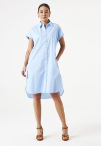 Hellblauses Hemdkleid mit kurzen Ärmeln, Knopfleiste, seitlichen Taschen und abgerundetem Saum. Glattes Material mit einer frischen Optik. Braune Absatzsandalen.