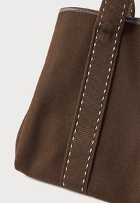 Καφέ σουέτ τσάντα χειρός με φαρδιά καφέ ραμμένη υφασμάτινη λαβή και λευκά ραμμένα αξεσουάρ σε απλό φόντο.