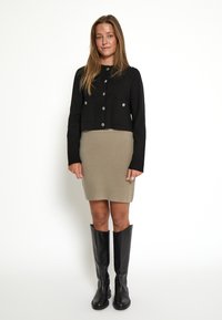 Giacca nera cropped con bottoni argentati, abbinata a un vestito beige fino al ginocchio e stivali neri al ginocchio, su uno sfondo neutro.