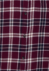 Camisa de franela a cuadros en rojo y marino que presenta un cierre de botones, una textura de tela suave y un bolsillo en el pecho con un detalle de botón negro.