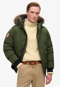 Grøn pufferjakke med en hætte af pels, lynlås foran og broderet mærke på ærmet; sat sammen med en cremet kabelstrikket sweater og beige bukser.