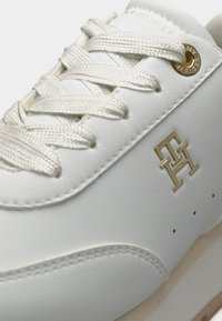 Άσπρο αθλητικό παπούτσι με υφασμένα κορδόνια, χρυσό λογότυπο Tommy Hilfiger στο πλάι και χρυσή τρύπα κορδονιού με σήμανση κοντά στην κορυφή.