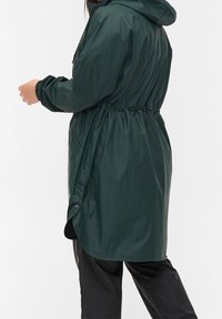 Mörkgrön vattentät parkas med huva, elastisk midja, tryckknappar och sidofickor. Slät, glansig textur, knälång design.
