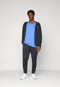Sort sportsjakke, blå t-shirt og grå sweatpants; hvide sneakers. Logo accenter på t-shirten og bukserne. Glat stofstruktur.
