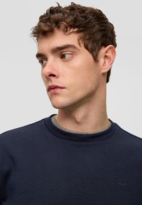 Felpa girocollo blu navy con tessuto a trama e un sottile logo "QS". Presenta polsini e scollatura a costine, indossata sopra una camicia grigia.