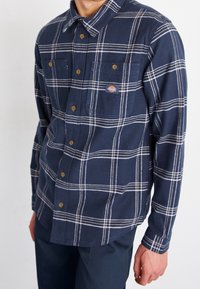 Camicia di flanella blu navy con motivo a quadri bianchi. Presenta due tasche sul petto, chiusura a bottoni e un colletto. Tessuto morbido.