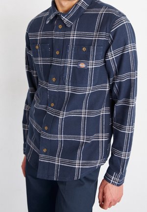 Chemise en flanelle bleu marine avec un motif à carreaux blancs. Comprend deux poches poitrine, une fermeture à boutons et un col. Texture douce.