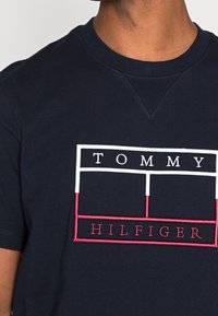 Tmavě modré bavlněné tričko s vyšívaným logem v bílé a červené barvě, které obsahuje obdélníkové vzory a zobrazuje text "TOMMY HILFIGER."