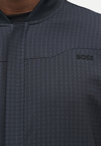 Κοντινή λήψη σκούρου, υφασματικού μπουφάν με φερμουάρ, ριμπ γιακά και ανάγλυφο λογότυπο "BOSS" στο στήθος.