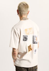Kurzes, cremefarbenes T-Shirt mit bunten Grafiken auf dem Rücken, das vielfältige Designs in Orange, Schwarz und Weiß zeigt.