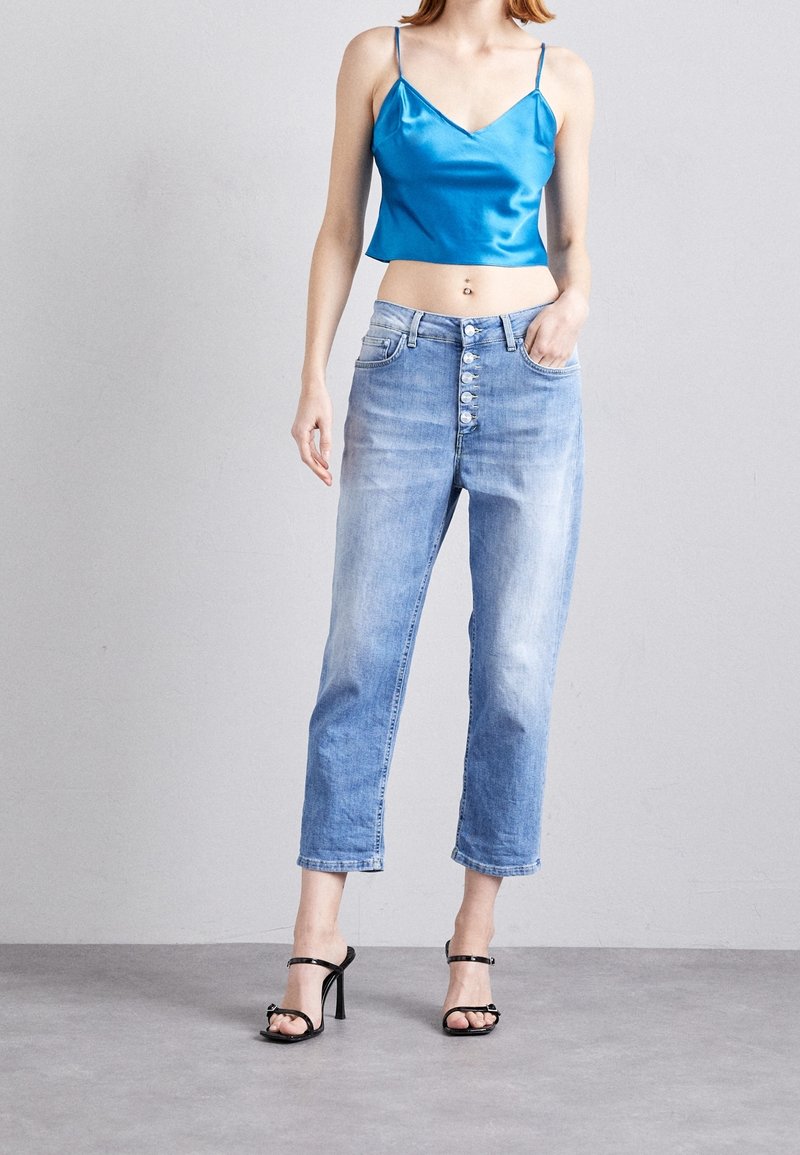 Tyrkysový saténový crop top s tenkými ramínky, zkombinovaný se světle modrými džínami s vysokým pasem, které mají knoflíkový zapínání a mírně zúžené nohavice. Černé sandály na podpatku.