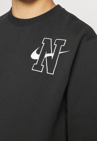 Černá bavlněná mikina s vyšívaným bílým logem „N“ a designem Nike Swoosh. Texturovaná látka s kulatým výstřihem.