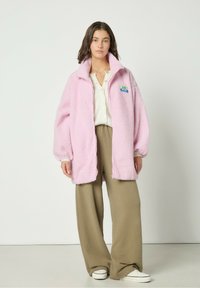 Roosa, tõmblukkudega, pehme jakk kõrge kaelusega ja logotäpiga. Kombineeritud beežide laia säärejäägiga pükstega ja valgete tossudega.