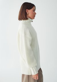 Maglione bianco lavorato a maglia con colletto alto a coste e vestibilità ampia, caratterizzato da motivi testurizzati e polsini a coste. Abbinato a pantaloni beige.