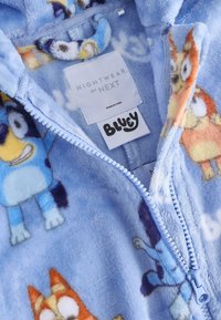 Vêtement de nuit en polaire bleu doux avec des personnages de chien dessin animé et une fermeture éclair à l'avant, étiqueté "Nightwear by Next" et étiquettes de marque "Bluey".