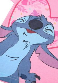 T-shirt rose clair avec un personnage de dessin animé à fourrure bleue, grandes oreilles et expression souriante. Design simple avec des accents floraux.