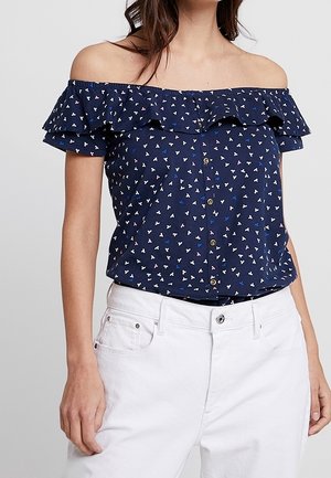 Mujer con blusa azul marino de volantes y hombros descubiertos, con pequeños patrones de corazones blancos y azules, combinada con pantalones blancos de talle alto.