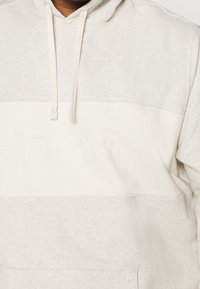 Helle beige Kapuzenjacke mit einem weißen horizontalen Einsatz und subtiler, tonaler "M STRAUSS & CO." Stickerei auf der Brust, ausgestattet mit Zugbändern an der Kapuze.