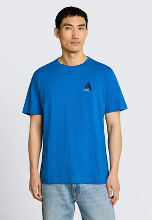 Mężczyzna noszący jednolitą, jasnoniebieską koszulkę z krótkim rękawem z małym logo żaglówki na lewej piersi, w połączeniu z jasnoniebieskimi dżinsami.