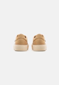 Tan fabric sneakers with a textured finish, featuring a thick woven sole and open heel design. Back view shows contouring details.