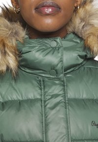 Gros plan d'une personne portant une doudoune verte avec une capuche bordée de fourrure et des boucles d'oreilles en forme de créoles dorées, lèvres fermées et regardant droit devant.