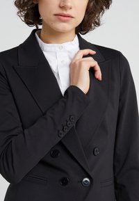 Blazer nero doppiopetto con rever a lancia, realizzato in tessuto liscio, con grandi bottoni neri e vestibilità su misura, indossato sopra una camicia bianca.