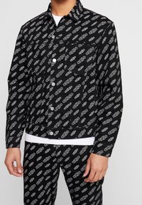 Svart denimjacka med vitt "CK50" logomönster, med knäppning framtill, spetsig krage och bröstficka. Texturen är slät.