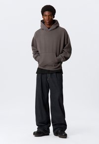 Grå hettegenser med frontlomme, kombinert med en svart lagdelt skjorte og løse mørke denimbukser. Svarte sko fullfører antrekket.